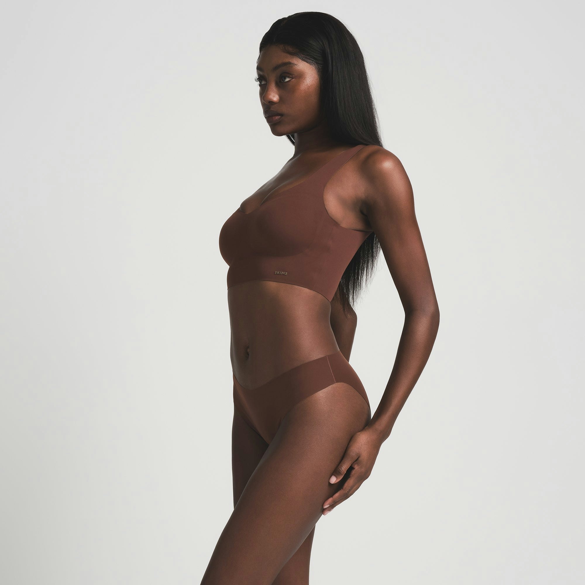 A MODEL STANDS FACING FORWARD WEARING THE SKIMS FREE CUT MID WAIST TANGA IN COCOA