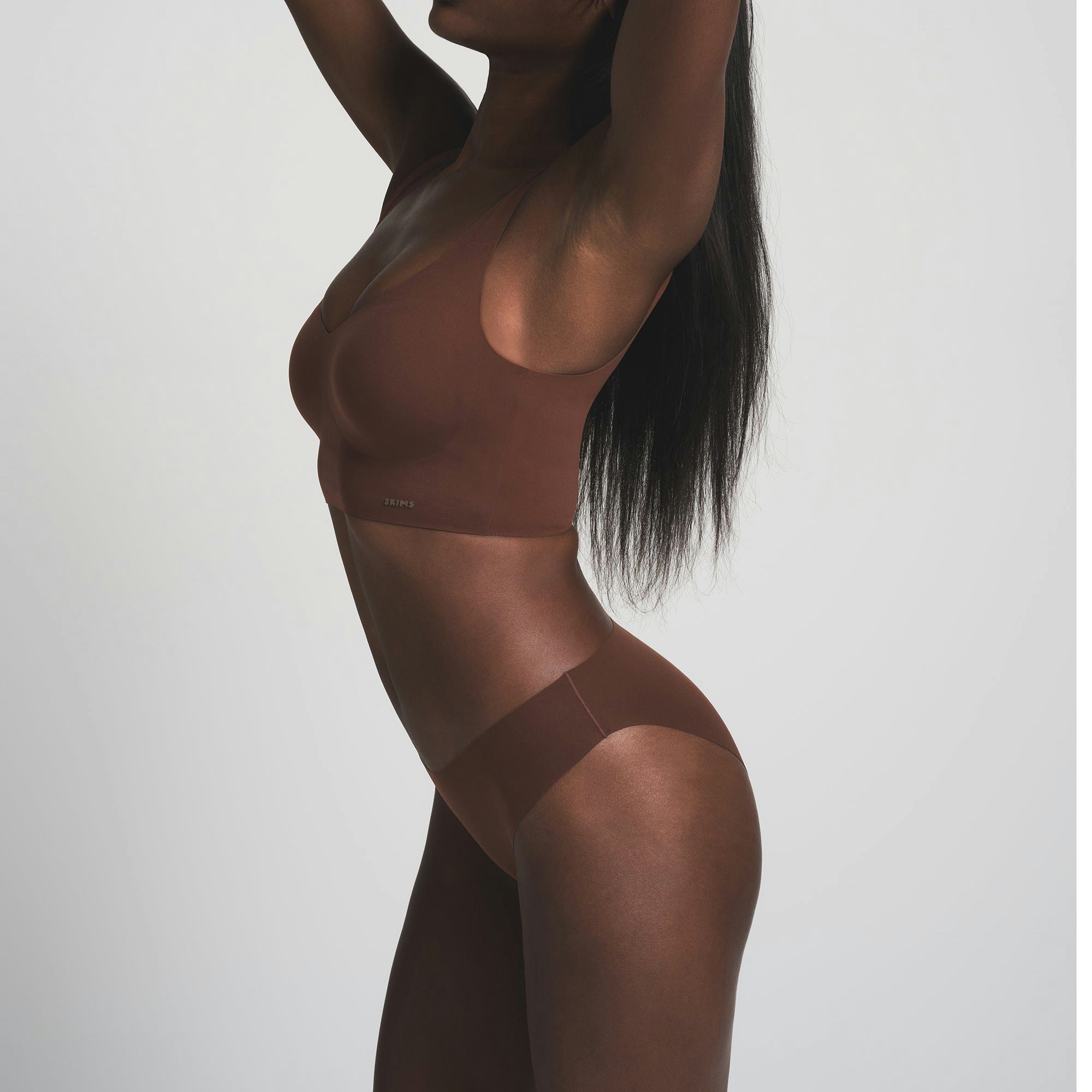 A MODEL STANDS FACING FORWARD WEARING THE SKIMS FREE CUT MID WAIST TANGA IN COCOA