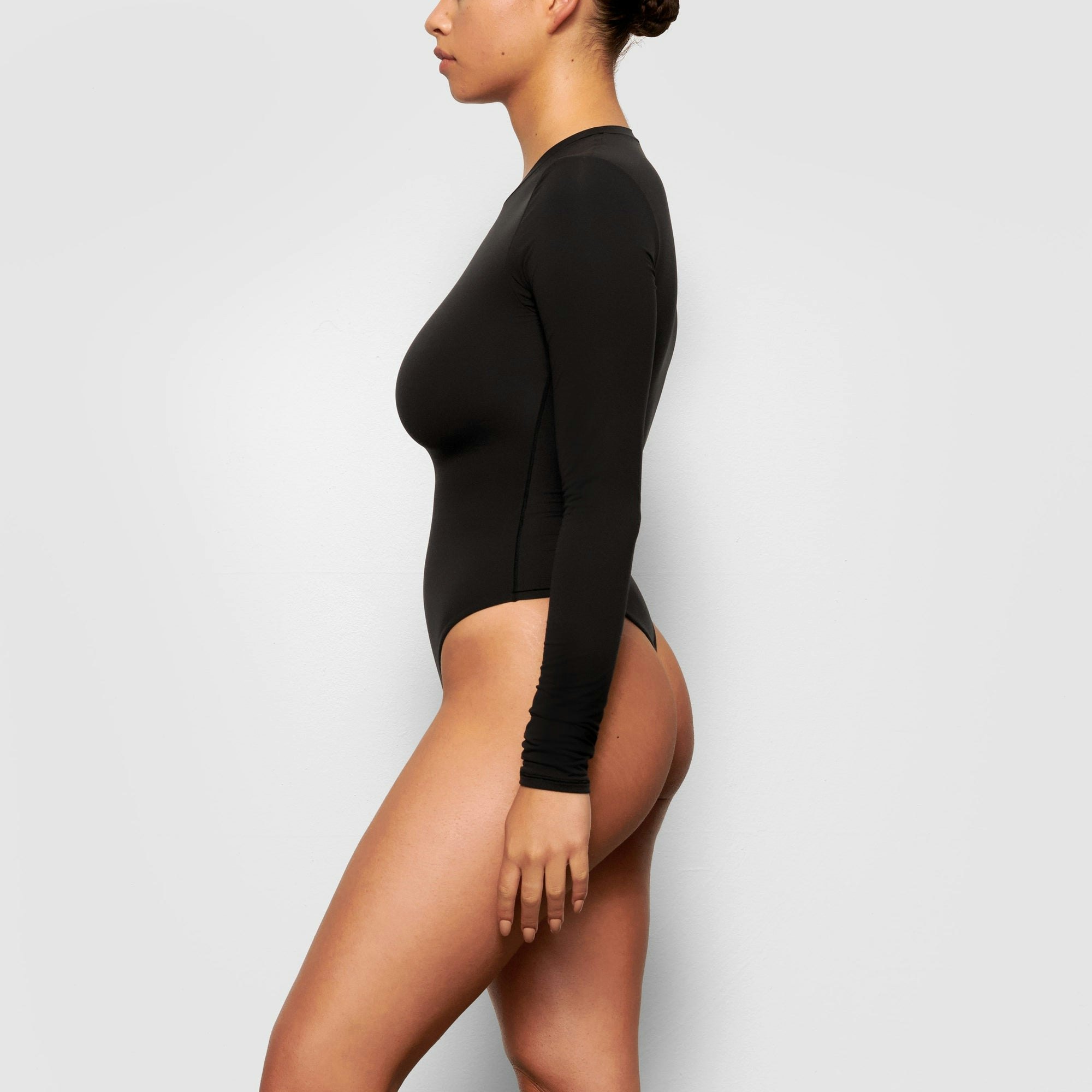 A MODEL STANDS SIDEWAYS SHOWING THE PROFILE OF THE FITS EVERYBODY LONG SLEEVE CREW NECK BODYSUIT | ONYX