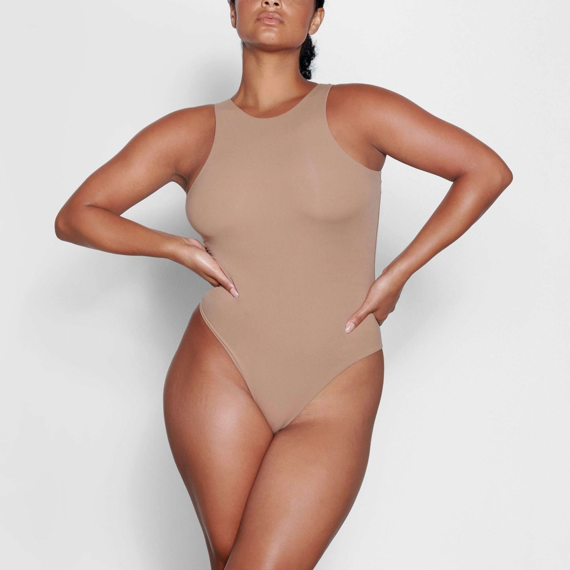A MODEL STANDS FACING FORWARD WEARING THE SKIMS FITS EVERYBODY HIGH NECK BODYSUIT IN SIENNA | SEE: M