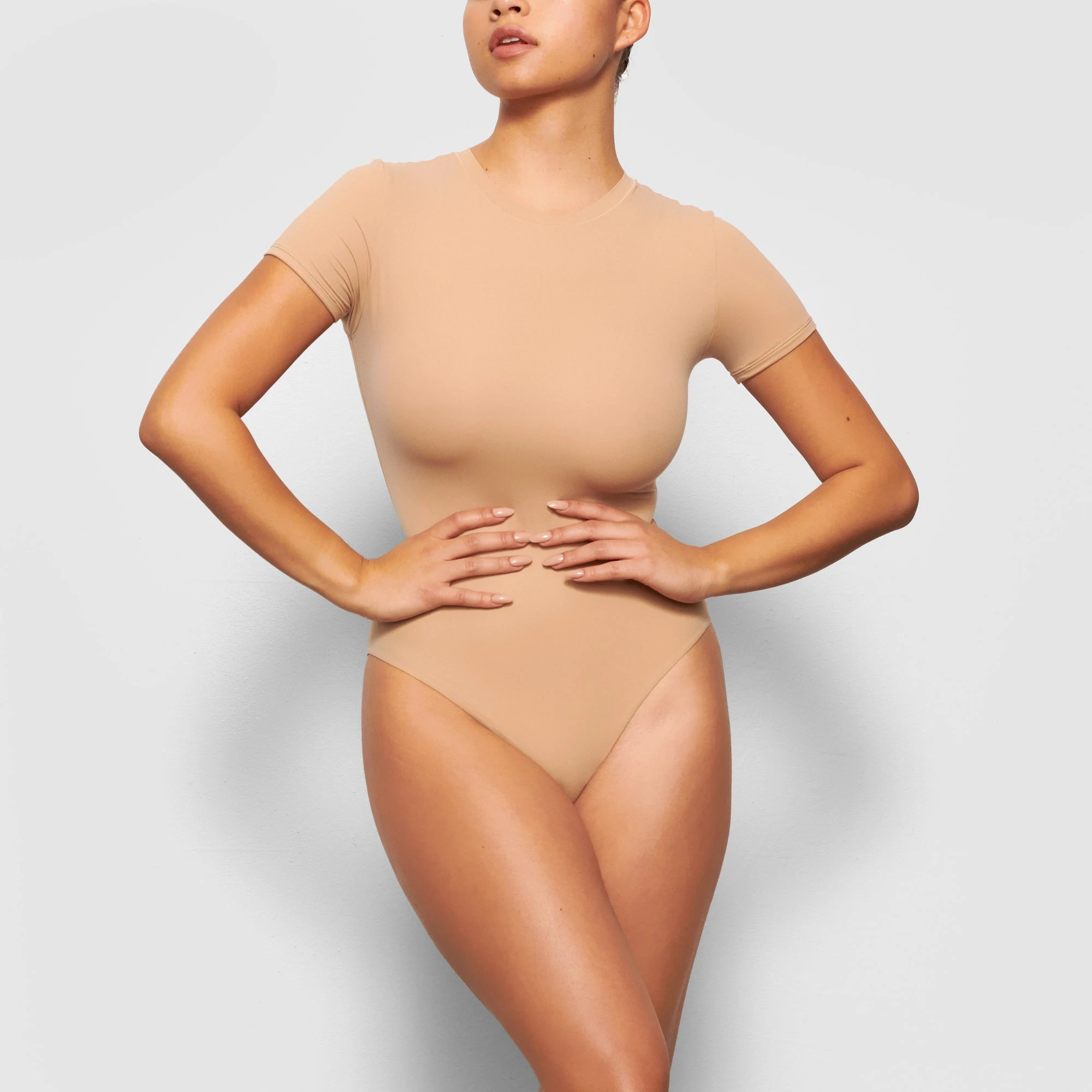 A MODEL STANDS FACING FORWARD WEARING THE SKIMS FITS EVERYBODY T-SHIRT BODYSUIT IN CLAY | SEE: XXS, XS, S