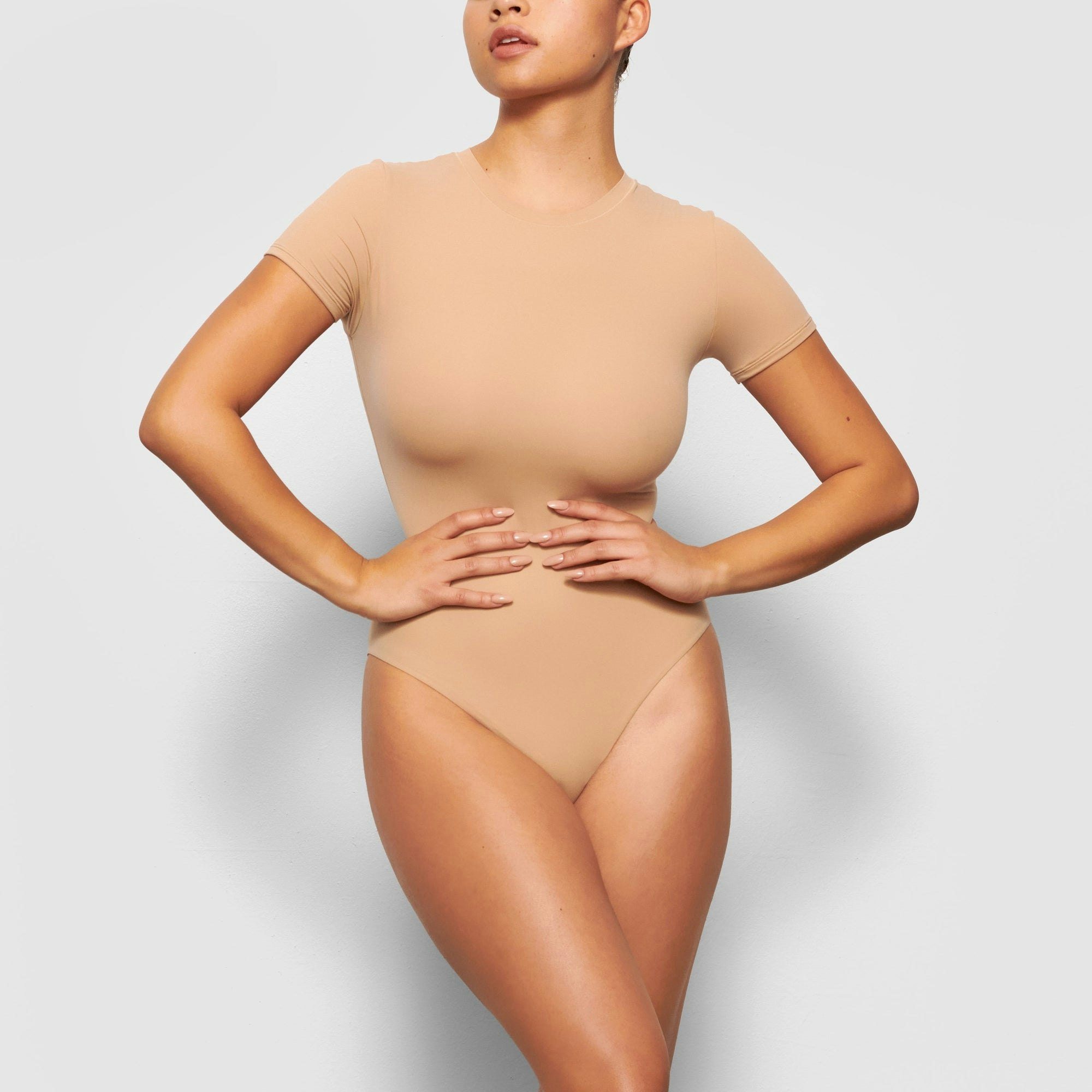 A MODEL STANDS FACING FORWARD WEARING THE SKIMS FITS EVERYBODY T-SHIRT BODYSUIT IN CLAY | SEE: XXS, XS, S