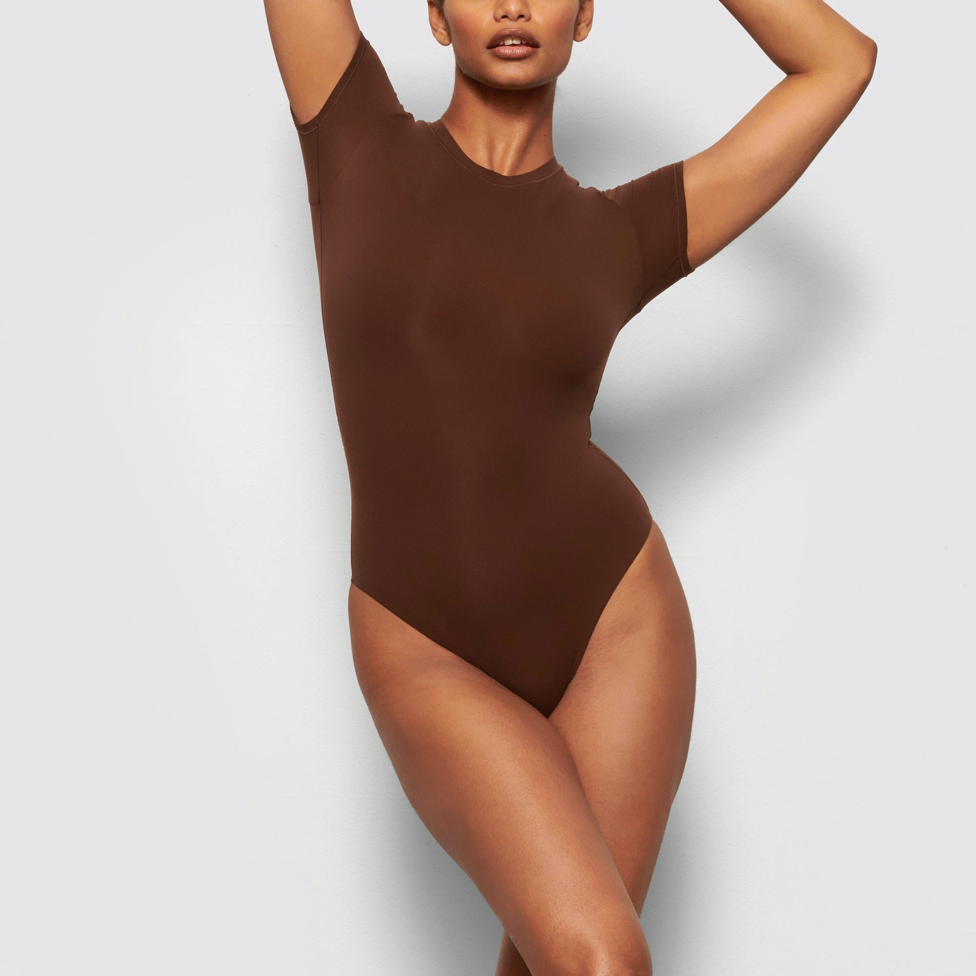 A MODEL STANDS FACING FORWARD WEARING THE SKIMS FITS EVERYBODY T-SHIRT BODYSUIT IN COCOA | SEE: M, L, XL