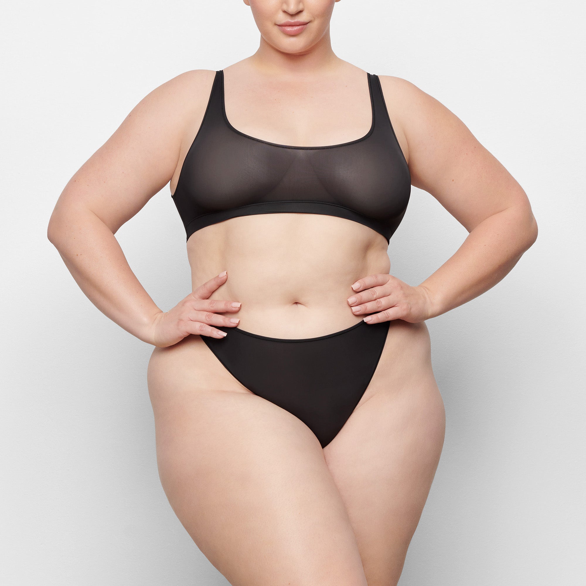 A MODEL STANDS FACING FORWARD WEARING THE SKIMS JELLY SHEER SCOOP NECK BRALETTE IN ONYX | SEE: 2X, 3X, 4X