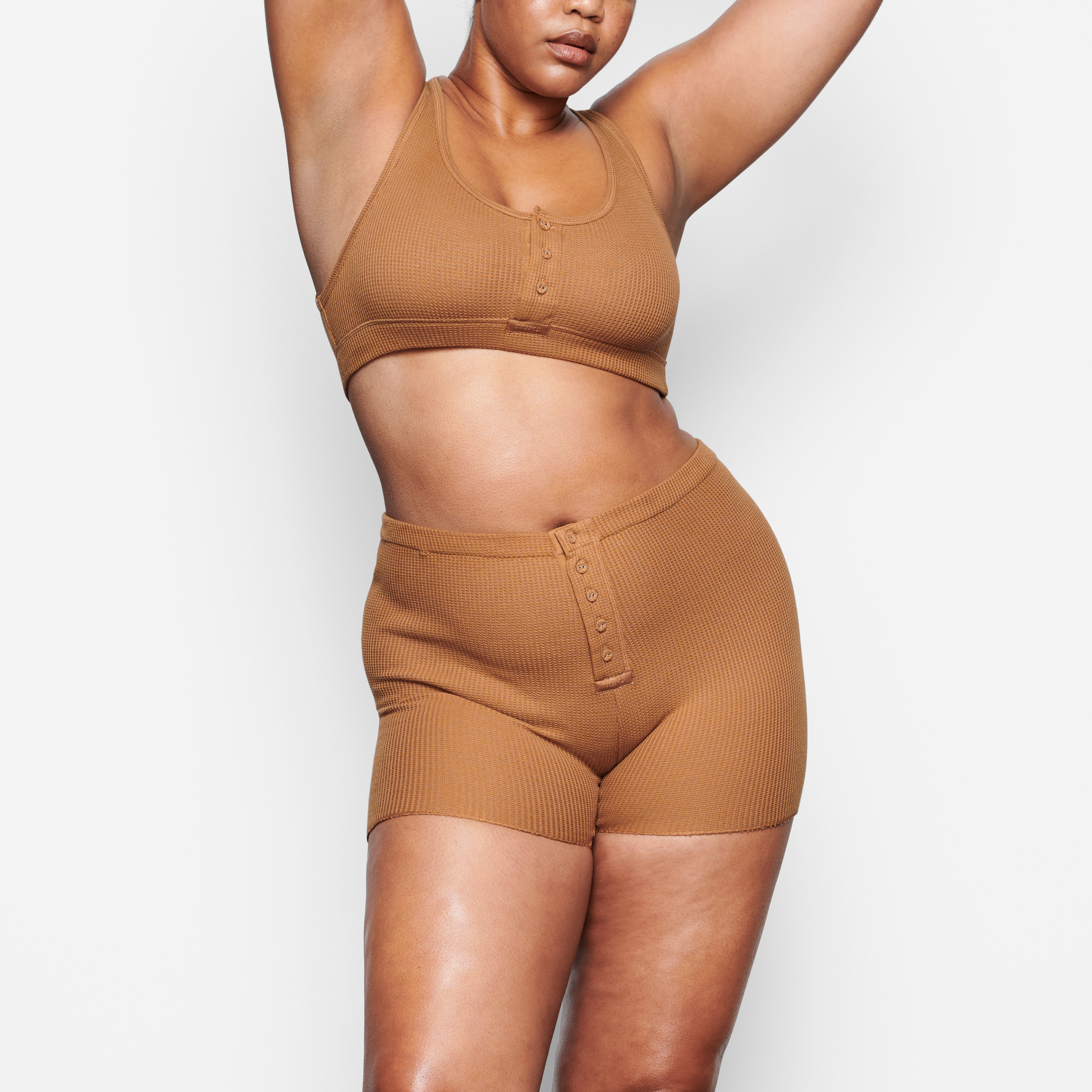 A MODEL STANDS FACING FORWARD WEARING THE SKIMS WAFFLE HENLEY BRA IN CAMEL | SEE: M, L, XL