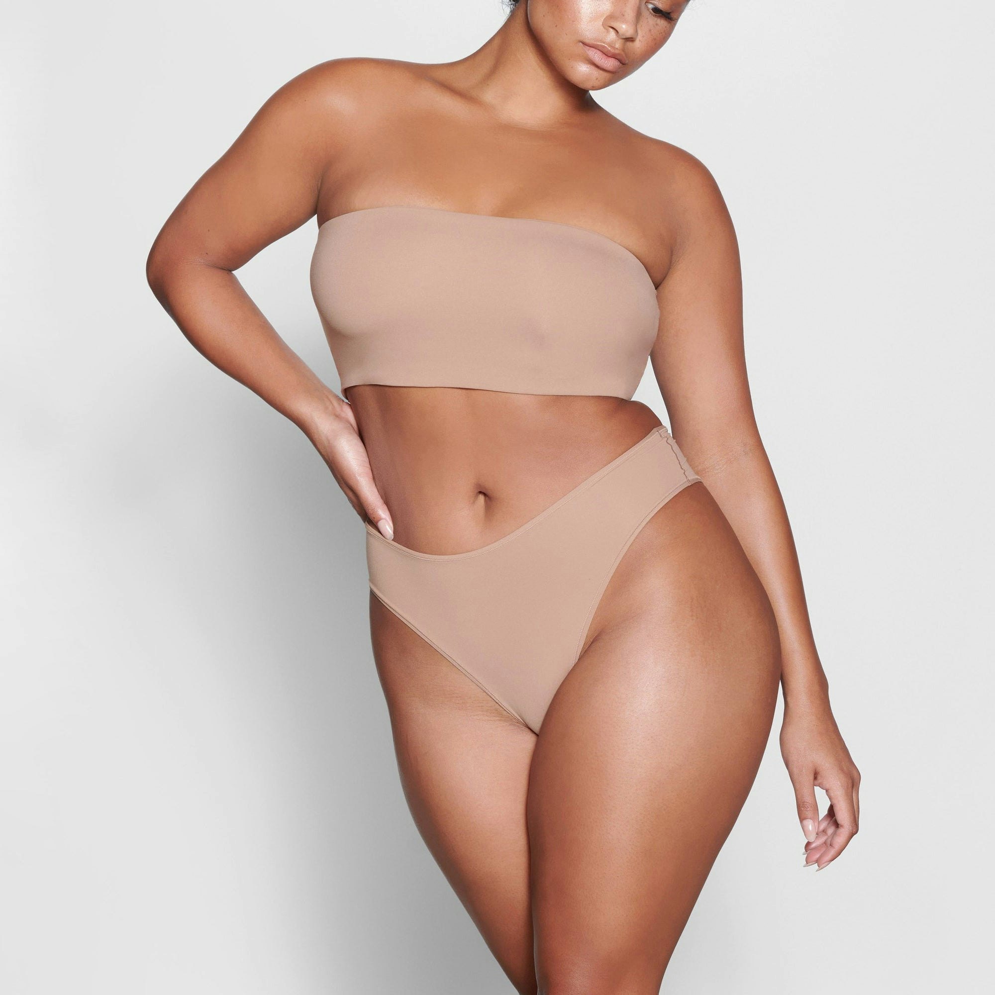 A MODEL STANDS FACING FORWARD WEARING THE SKIMS FITS EVERYBODY BANDEAU BRA IN SIENNA | SEE: M