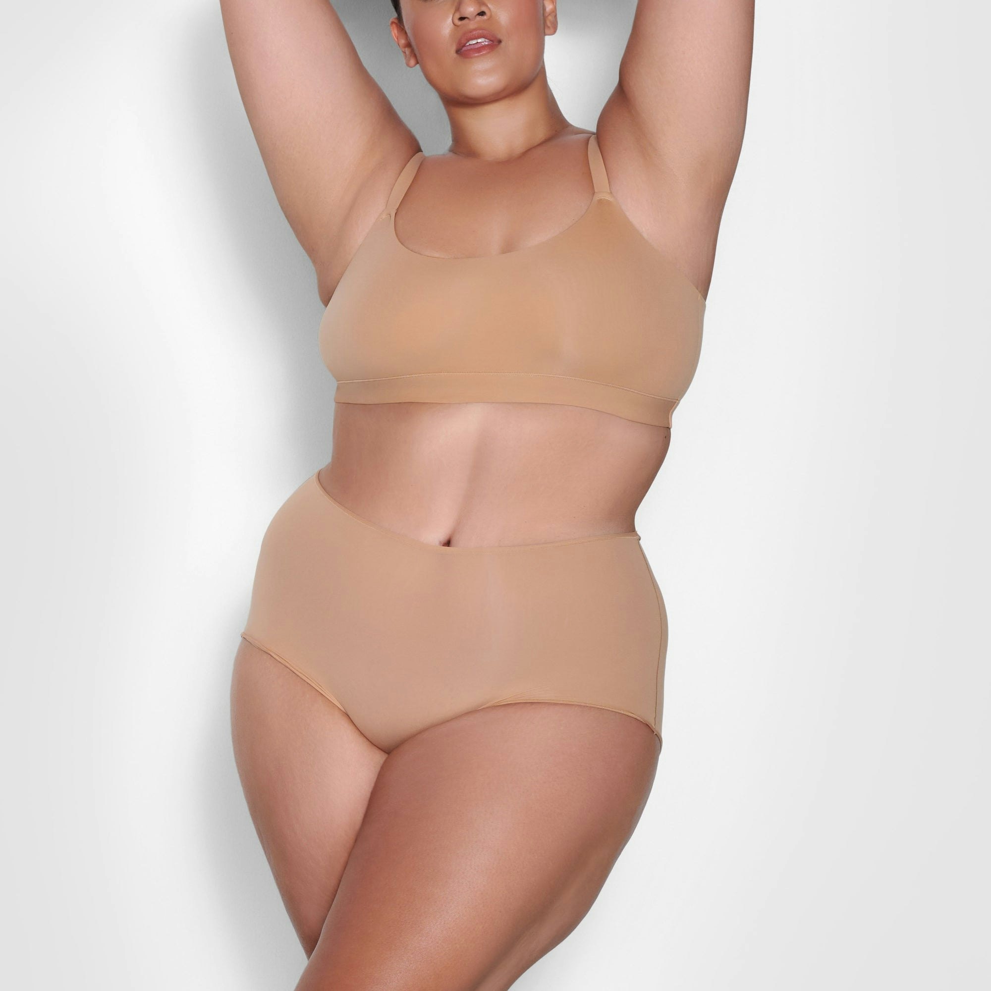 A MODEL STANDS FACING FORWARD WEARING THE SKIMS FITS EVERYBODY SCOOP NECK BRA IN OCHRE | SEE: 2X, 3X, 4X