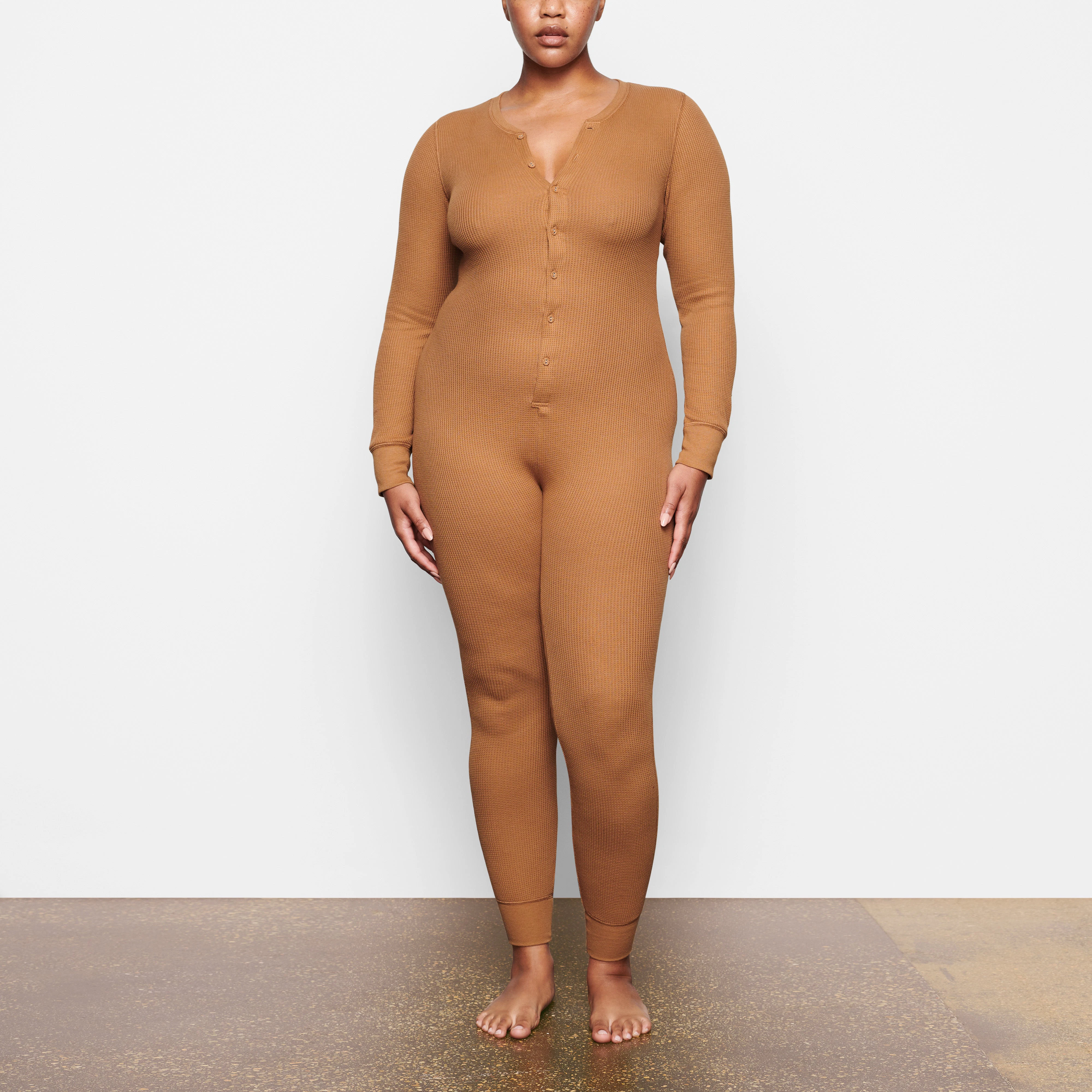 A MODEL STANDS FACING FORWARD WEARING THE SKIMS WAFFLE ONESIE IN CAMEL | SEE: M, L, XL