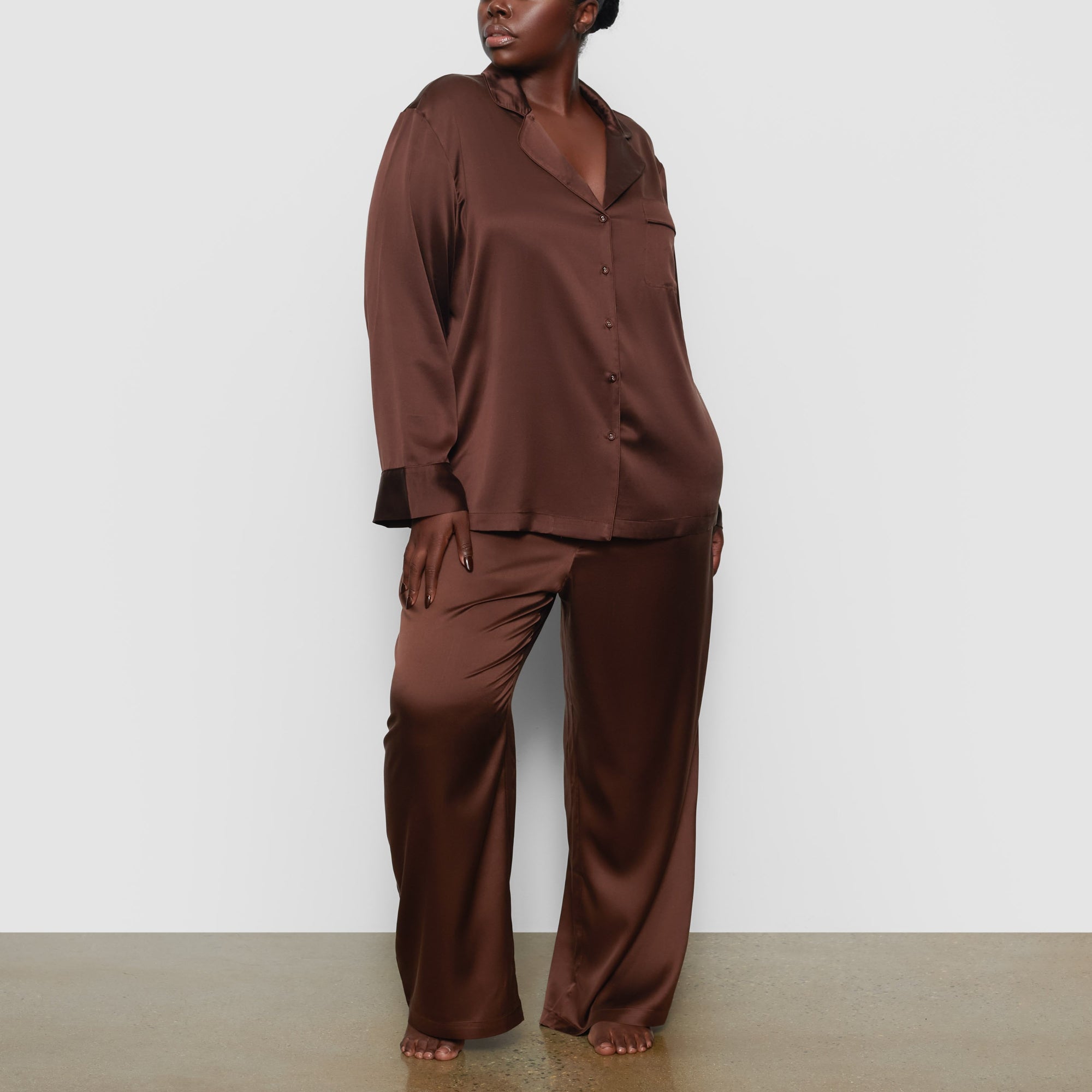 A MODEL STANDS FACING FORWARD WEARING THE SKIMS SILK BUTTON UP SHIRT IN COCOA | SEE: 2X, 3X, 4X