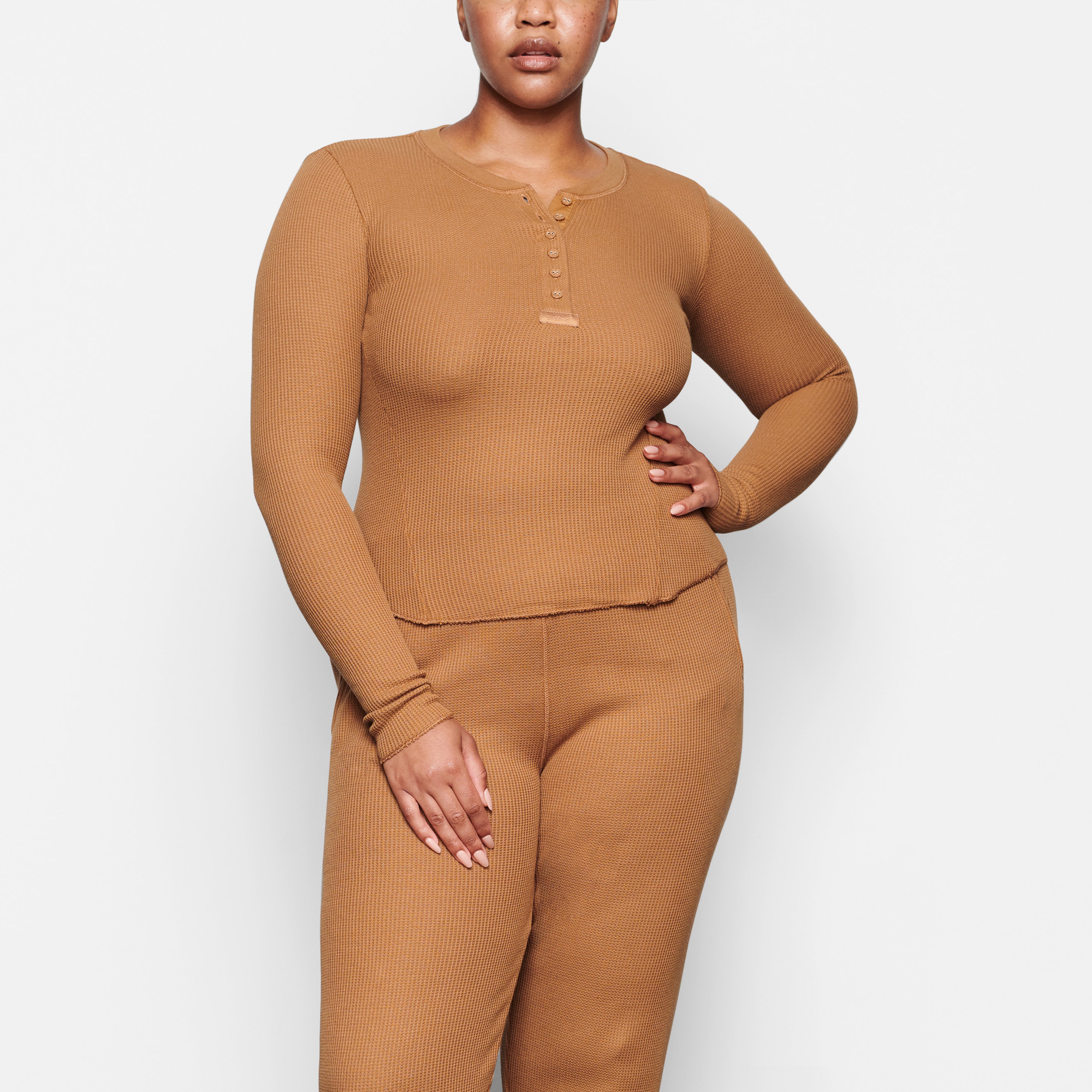 A MODEL STANDS FACING FORWARD WEARING THE SKIMS WAFFLE LONG SLEEVE HENLEY IN CAMEL | SEE: M, L, XL