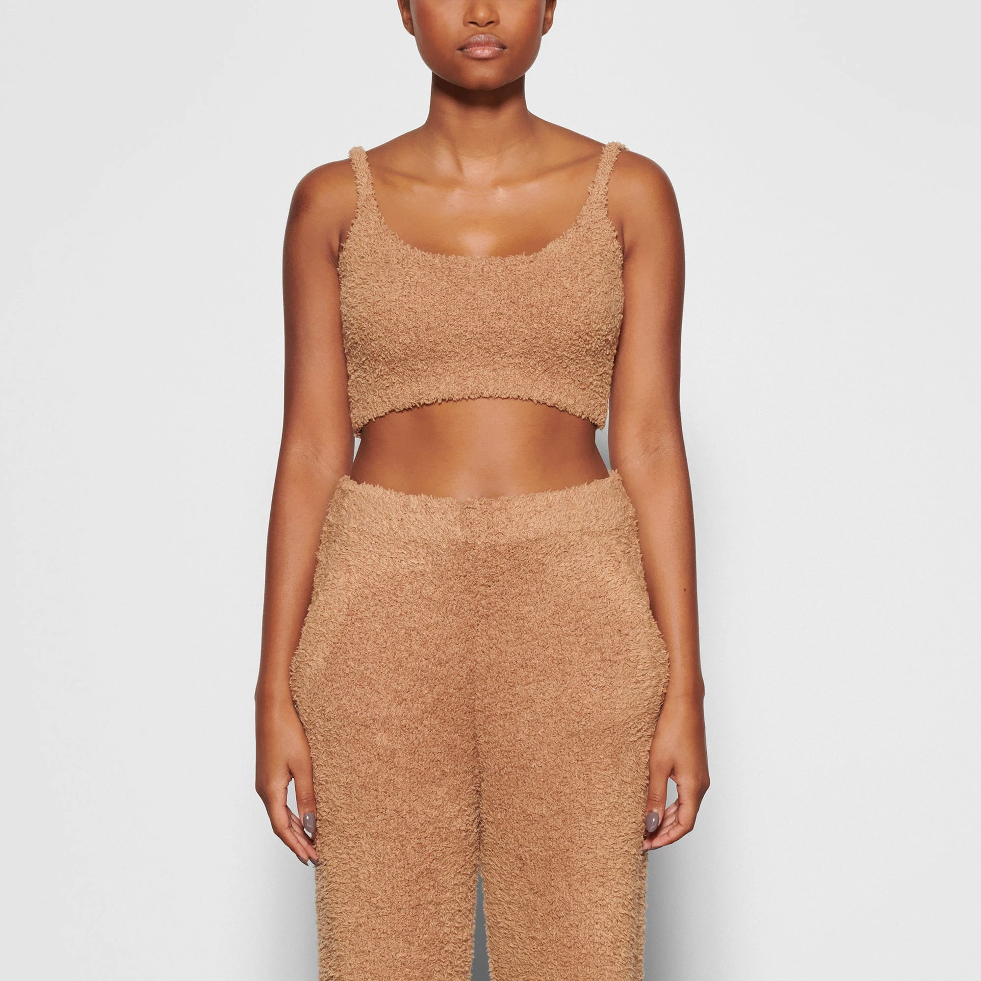 A MODEL STANDS FACING FORWARD WEARING THE SKIMS COZY KNIT BRALETTE IN CAMEL | SEE: XXS/XS
