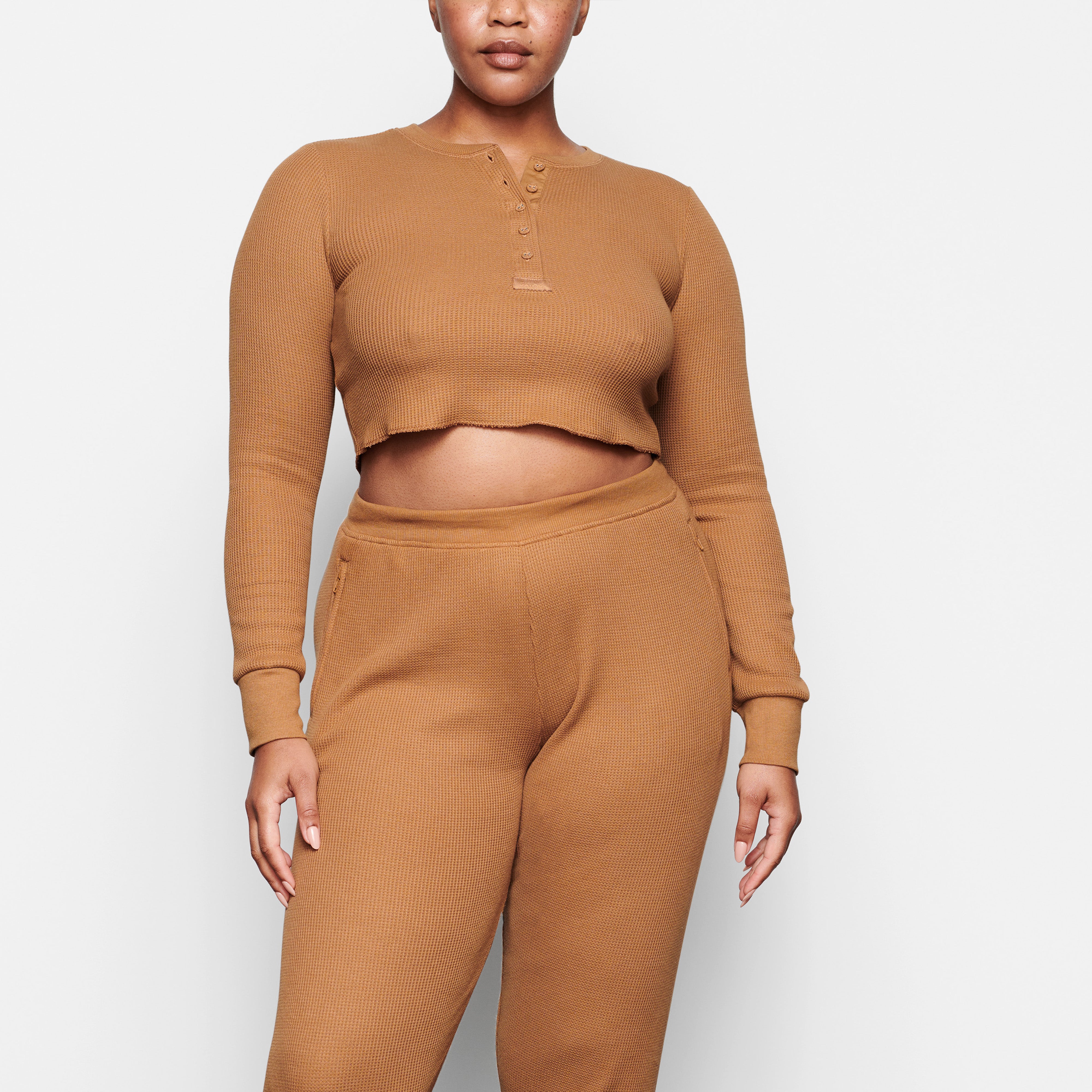 A MODEL STANDS FACING FORWARD WEARING THE SKIMS WAFFLE LONG SLEEVE CROP HENLEY IN CAMEL | SEE: M, L, XL