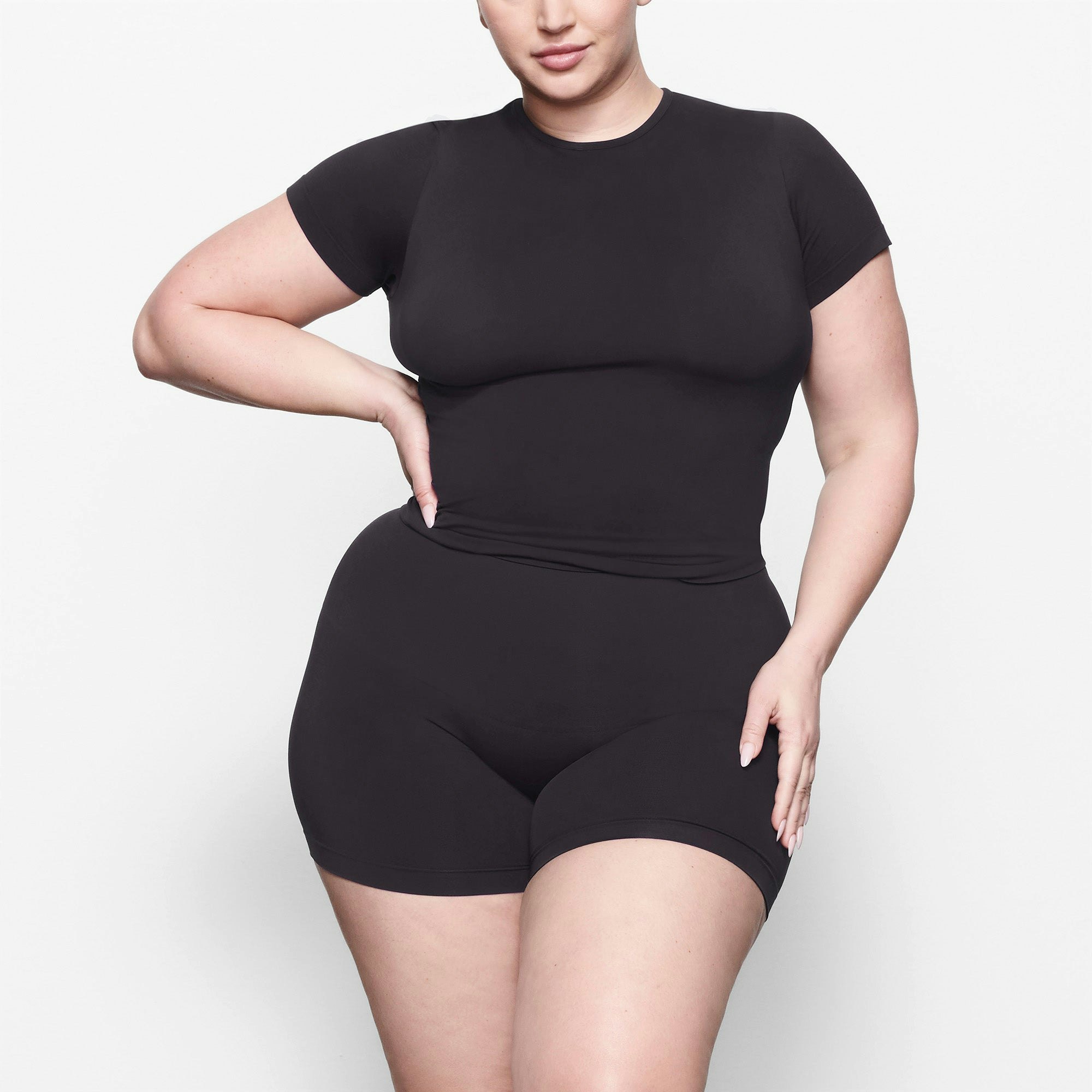 A MODEL STANDS FACING FORWARD WEARING THE SKIMS SOFT SMOOTHING T-SHIRT IN ONYX | SEE: 2X, 3X, 4X