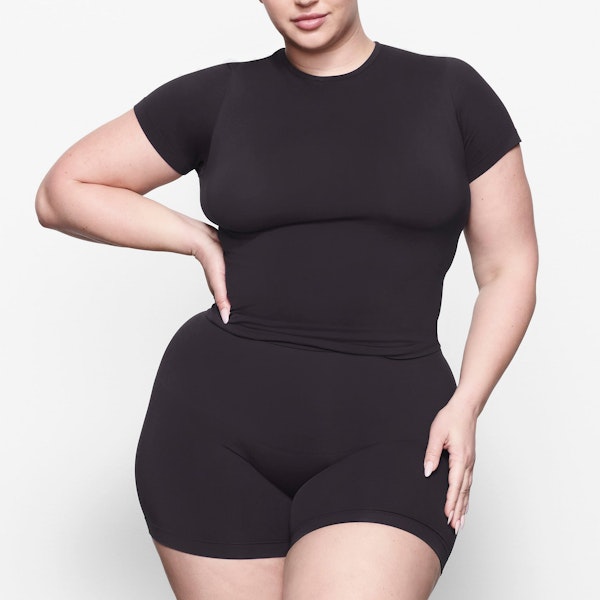 A MODEL STANDS FACING FORWARD WEARING THE SKIMS SOFT SMOOTHING T-SHIRT IN ONYX | SEE: 2X, 3X, 4X