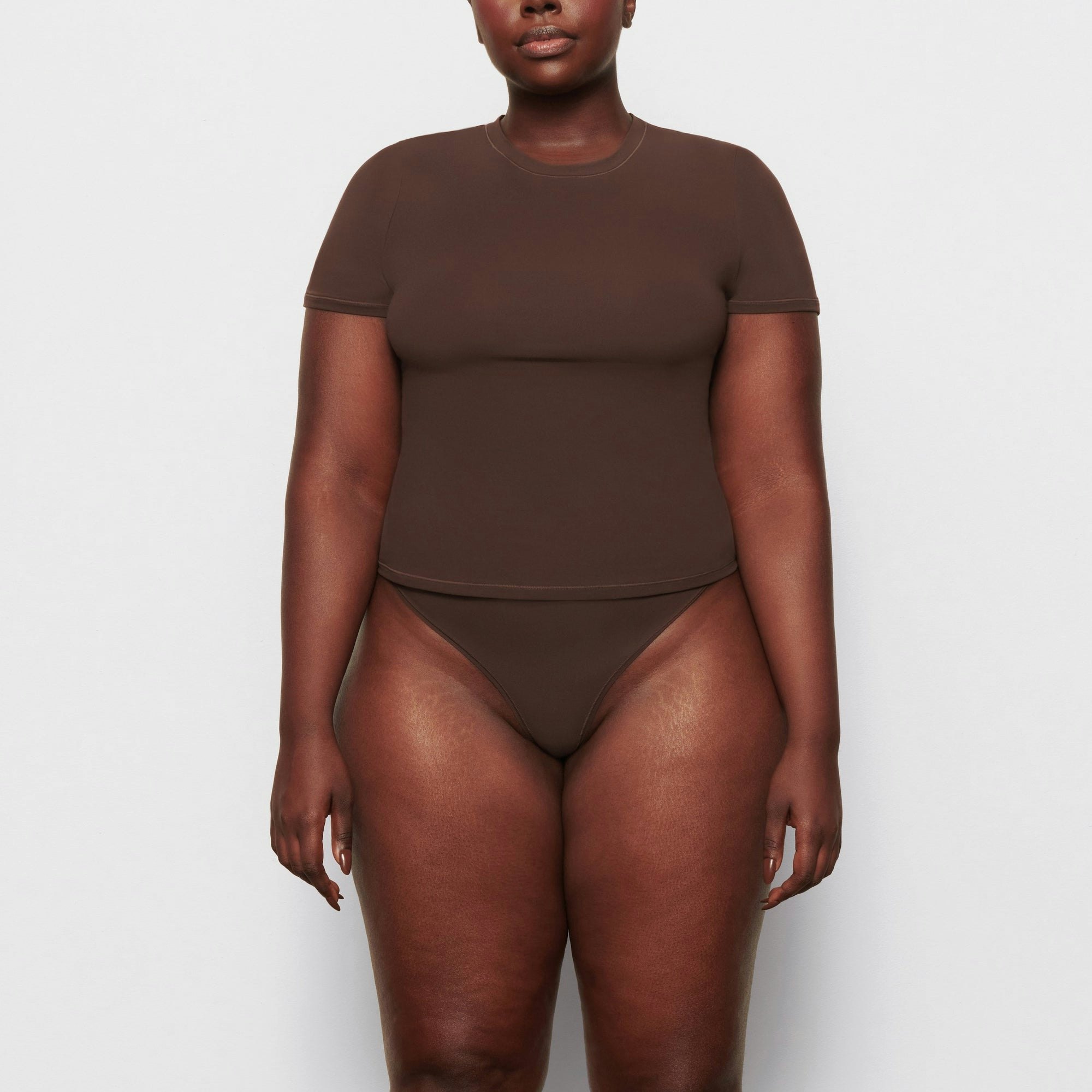 A MODEL STANDS FACING FORWARD WEARING THE SKIMS FITS EVERYBODY T-SHIRT IN COCOA