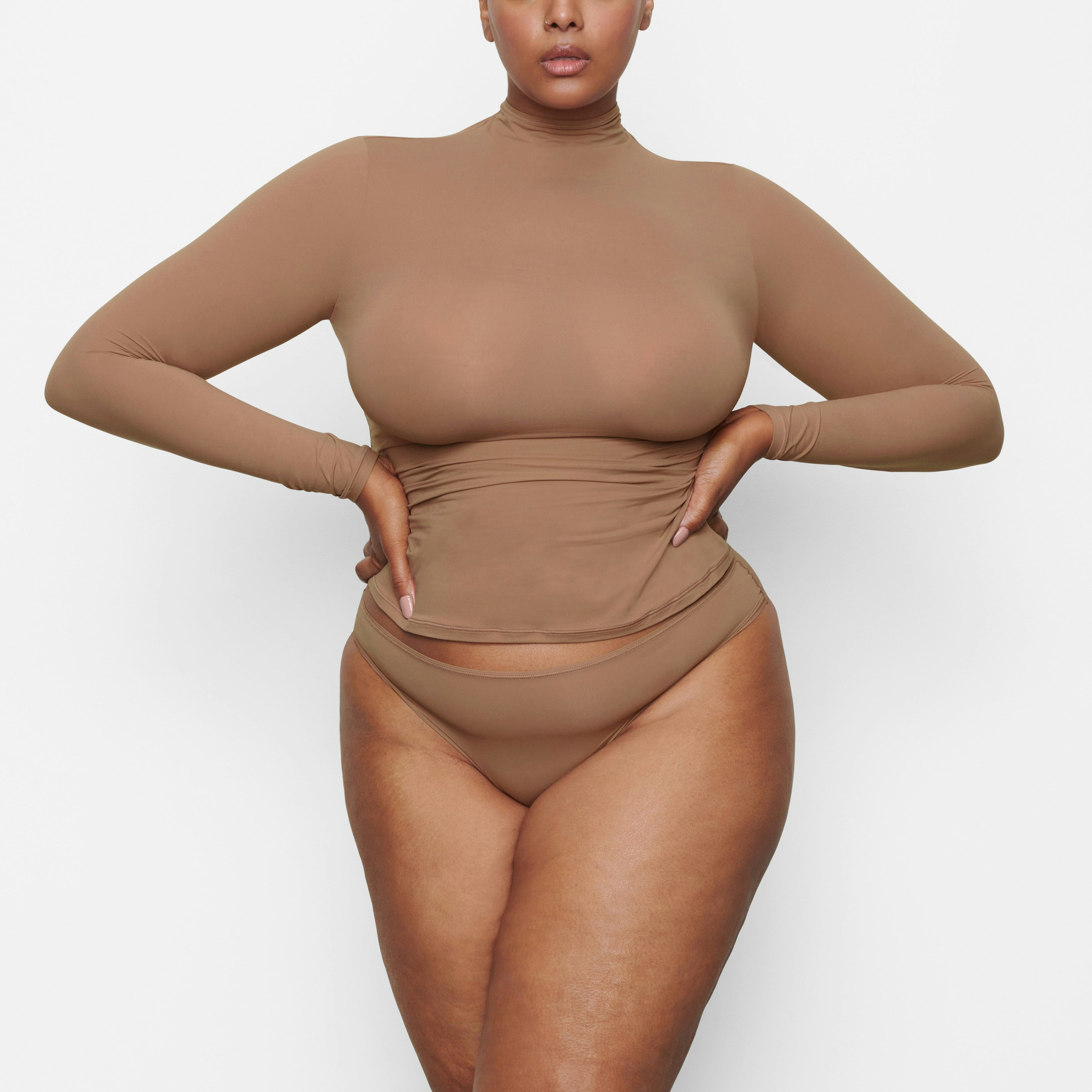 A MODEL STANDS FACING FORWARD WEARING THE SKIMS FITS EVERYBODY TURTLENECK TOP IN SIENNA