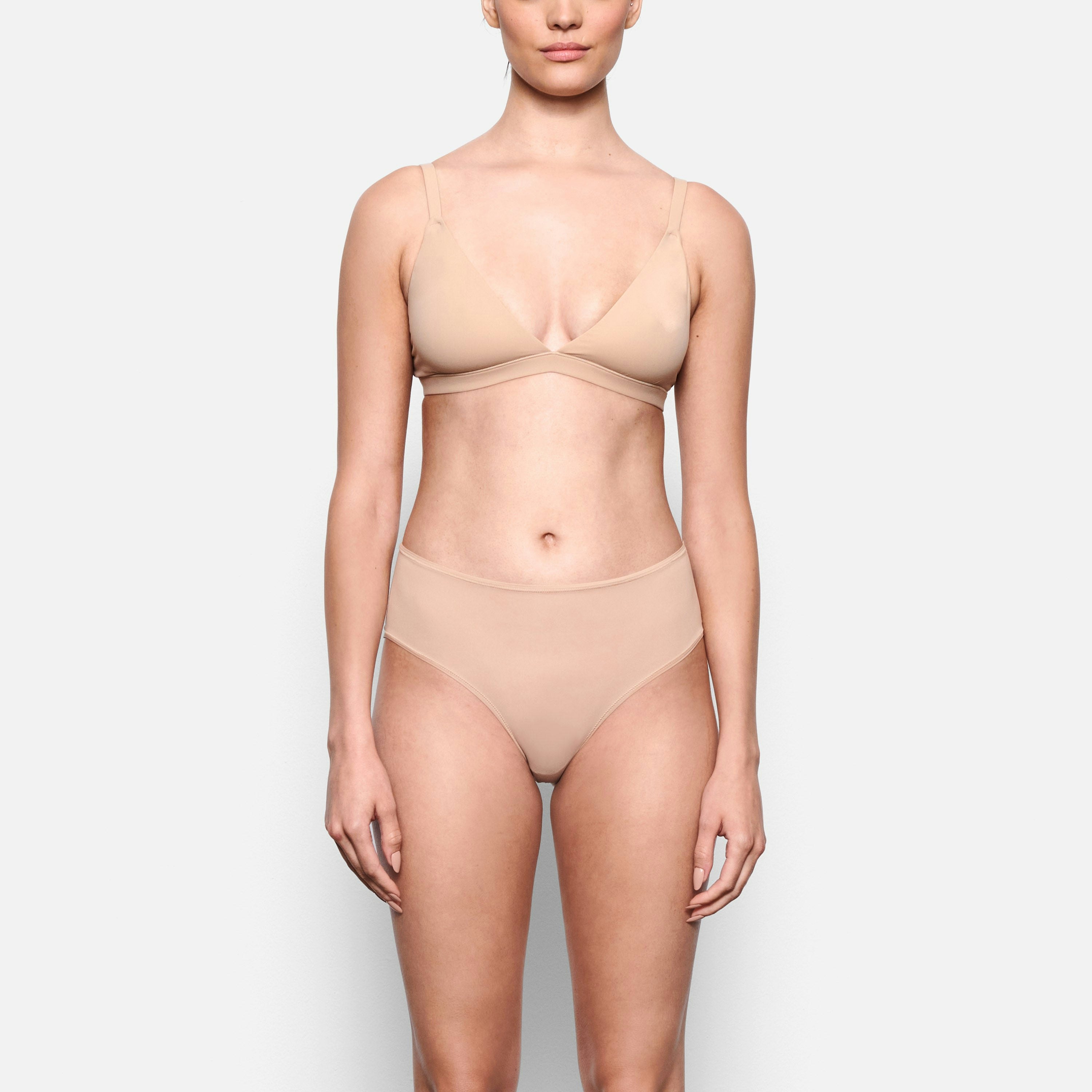A MODEL STANDS FACING FORWARD WEARING THE SKIMS FITS EVERYBODY MID RISE BRIEF IN CLAY | SEE: XXS, XS, S