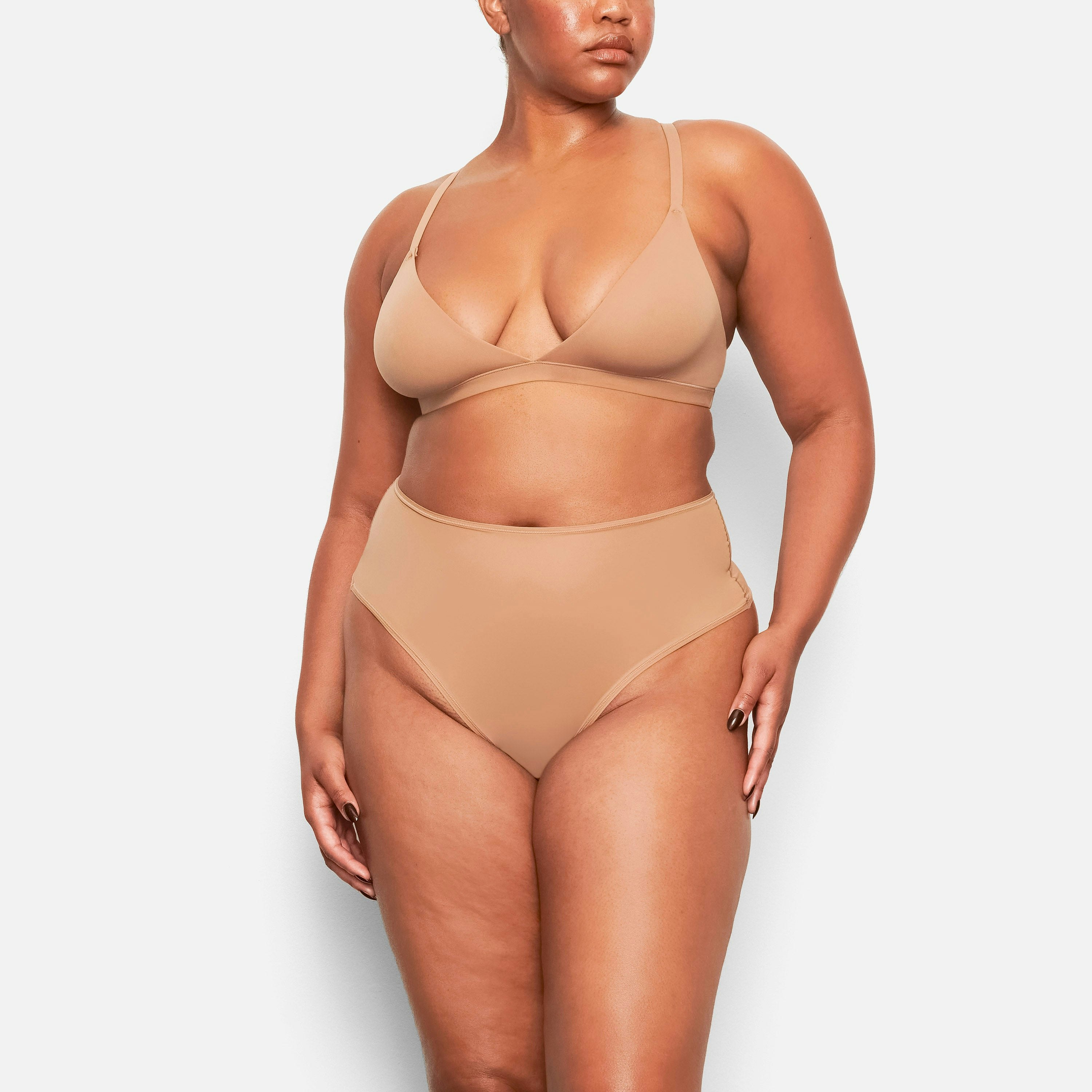 A MODEL STANDS FACING FORWARD WEARING THE SKIMS FITS EVERYBODY HIGH WAISTED THONG IN SIENNA | SEE: M, L, XL
