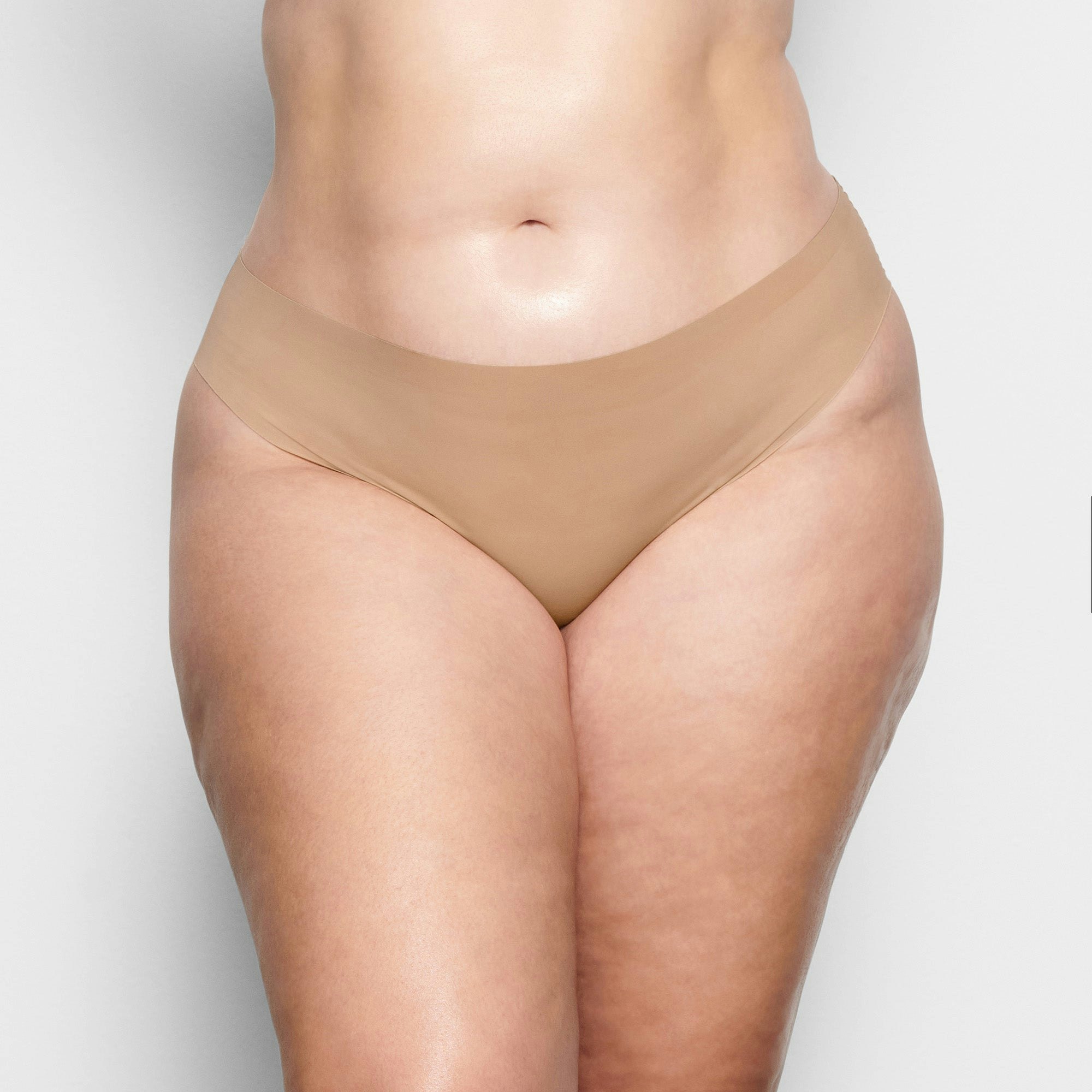 A MODEL STANDS FACING FORWARD WEARING THE SKIMS FREE CUT MID WAIST TANGA IN CLAY | SEE: 2X, 3X, 4X | FOCUS: 0.1, 0.9, 0.9