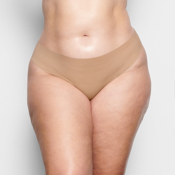 A MODEL STANDS FACING FORWARD WEARING THE SKIMS FREE CUT MID WAIST TANGA IN CLAY | SEE: 2X, 3X, 4X | FOCUS: 0.1, 0.9, 0.9