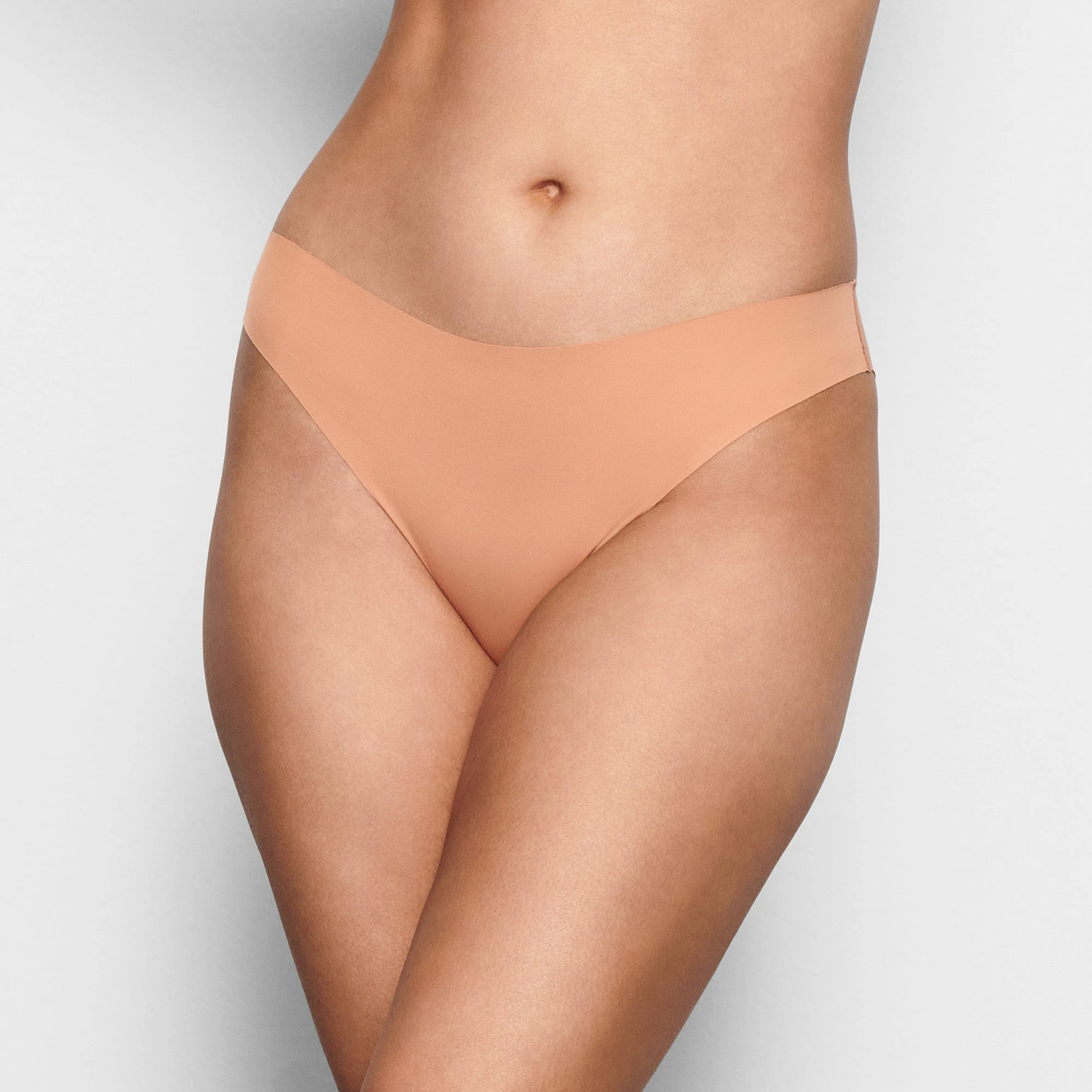 A MODEL STANDS FACING FORWARD WEARING THE SKIMS FREE CUT MID WAIST TANGA IN OCHRE | SEE: XXS, XS, S| FOCUS: 0.1, 0.9, 0.9