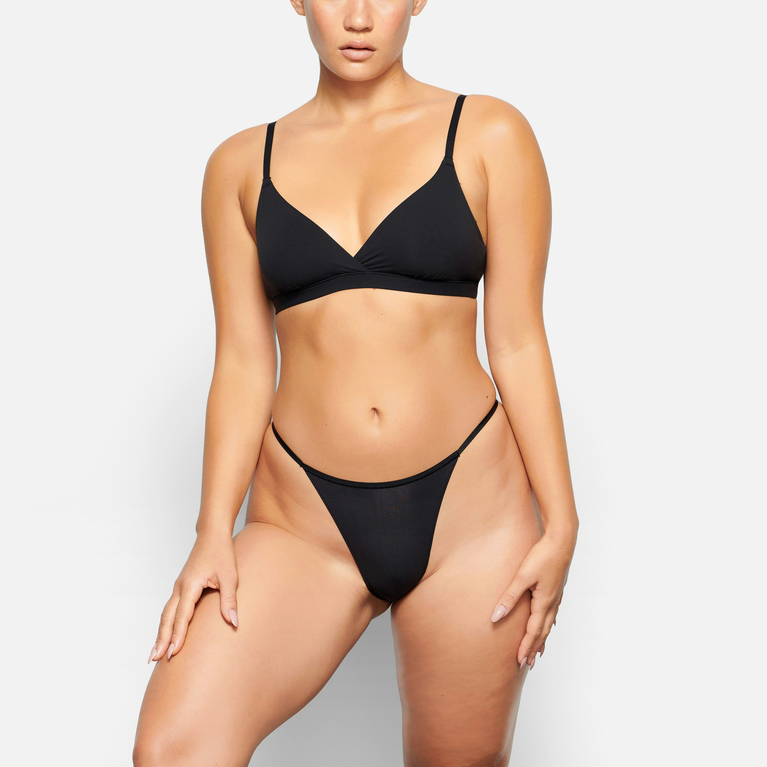 A MODEL STANDS FACING FORWARD WEARING THE SKIMS FITS EVERYBODY CHEEKY STRING BIKINI IN ONYX | SEE: XXS, XS, S