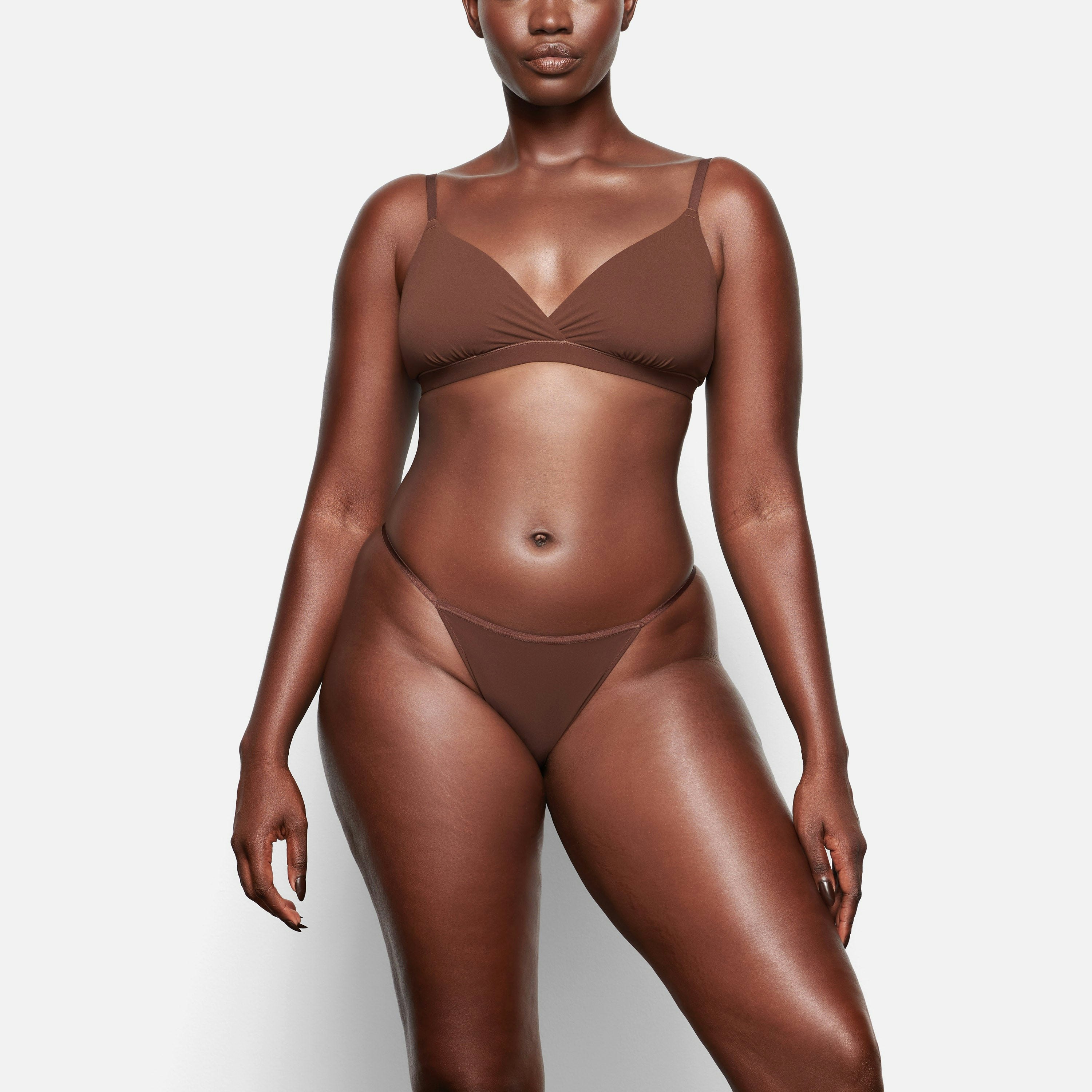 A MODEL STANDS FACING FORWARD WEARING THE SKIMS FITS EVERYBODY CHEEKY STRING BIKINI IN COCOA | SEE: M, L, XL