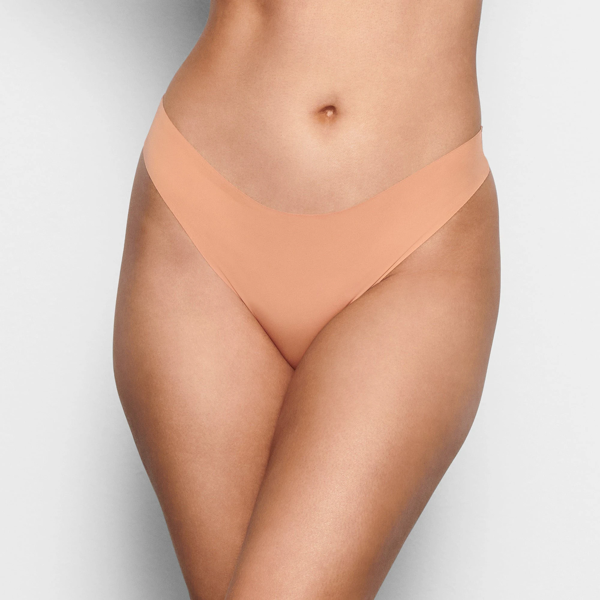 A MODEL STANDS FACING FORWARD WEARING THE SKIMS FREE CUT MID WAIST THONG IN OCHRE | SEE: XXS, XS, S| FOCUS: 0.1, 0.9, 0.9
