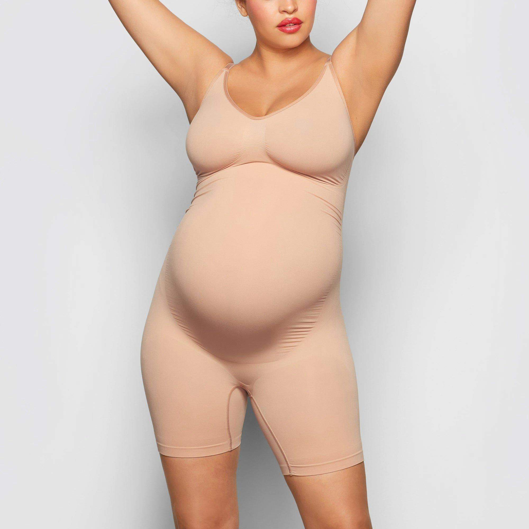 A MODEL STANDS FACING FORWARD WEARING THE SKIMS MATERNITY SCULPTING BODYSUIT MID THIGH IN SIENNA | SEE: S/M