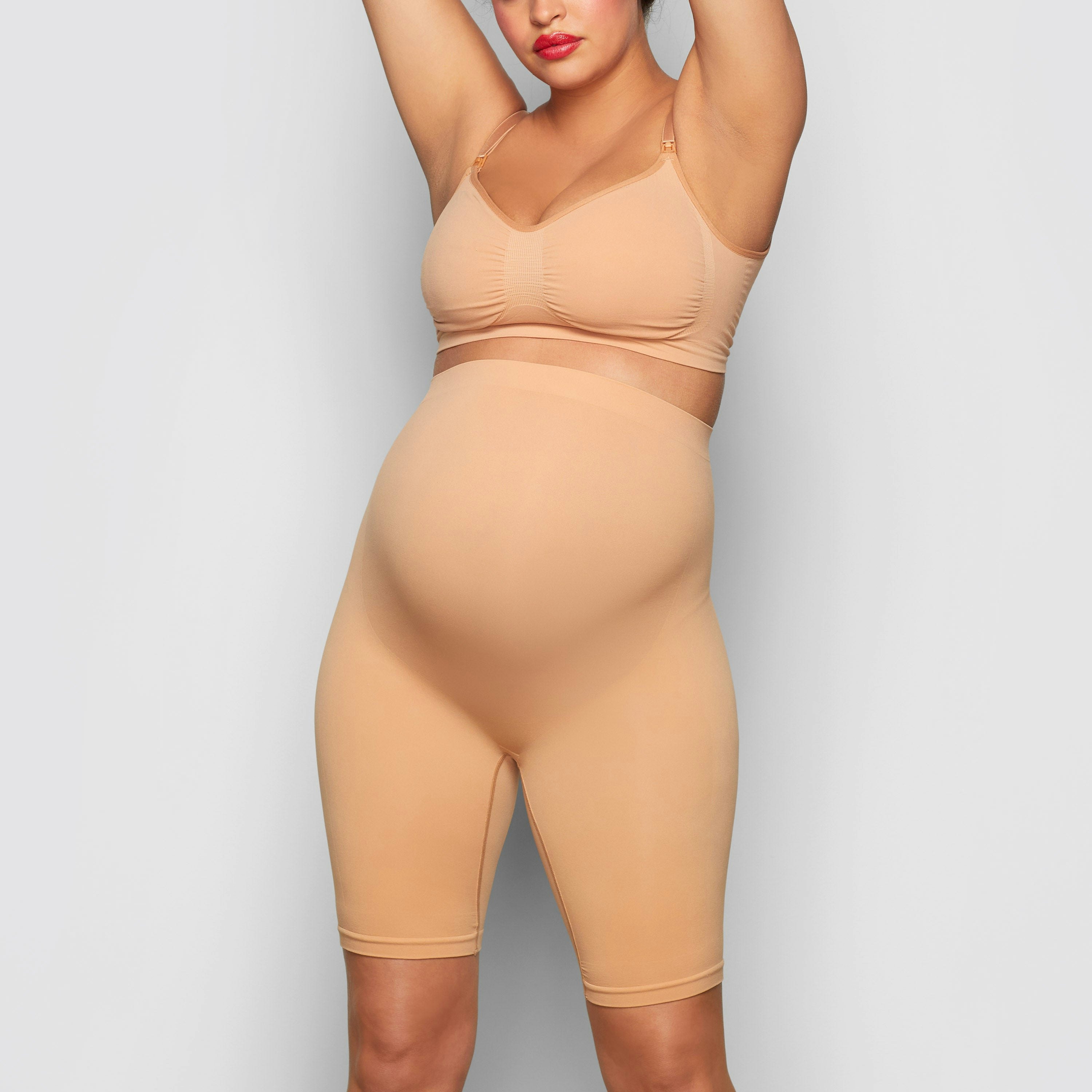 A MODEL STANDS FACING FORWARD WEARING THE SKIMS MATERNITY SCULPTING SHORT MID THIGH IN OCHRE | SEE: S/M