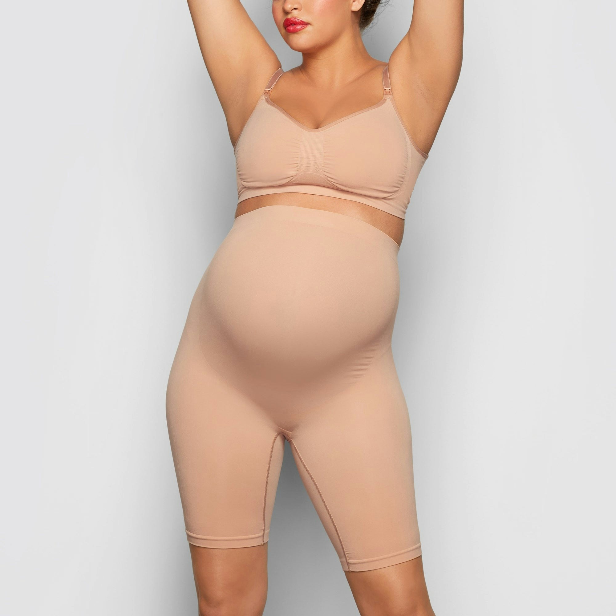 A MODEL STANDS FACING FORWARD WEARING THE SKIMS MATERNITY SCULPTING SHORT MID THIGH IN SIENNA | SEE: S/M