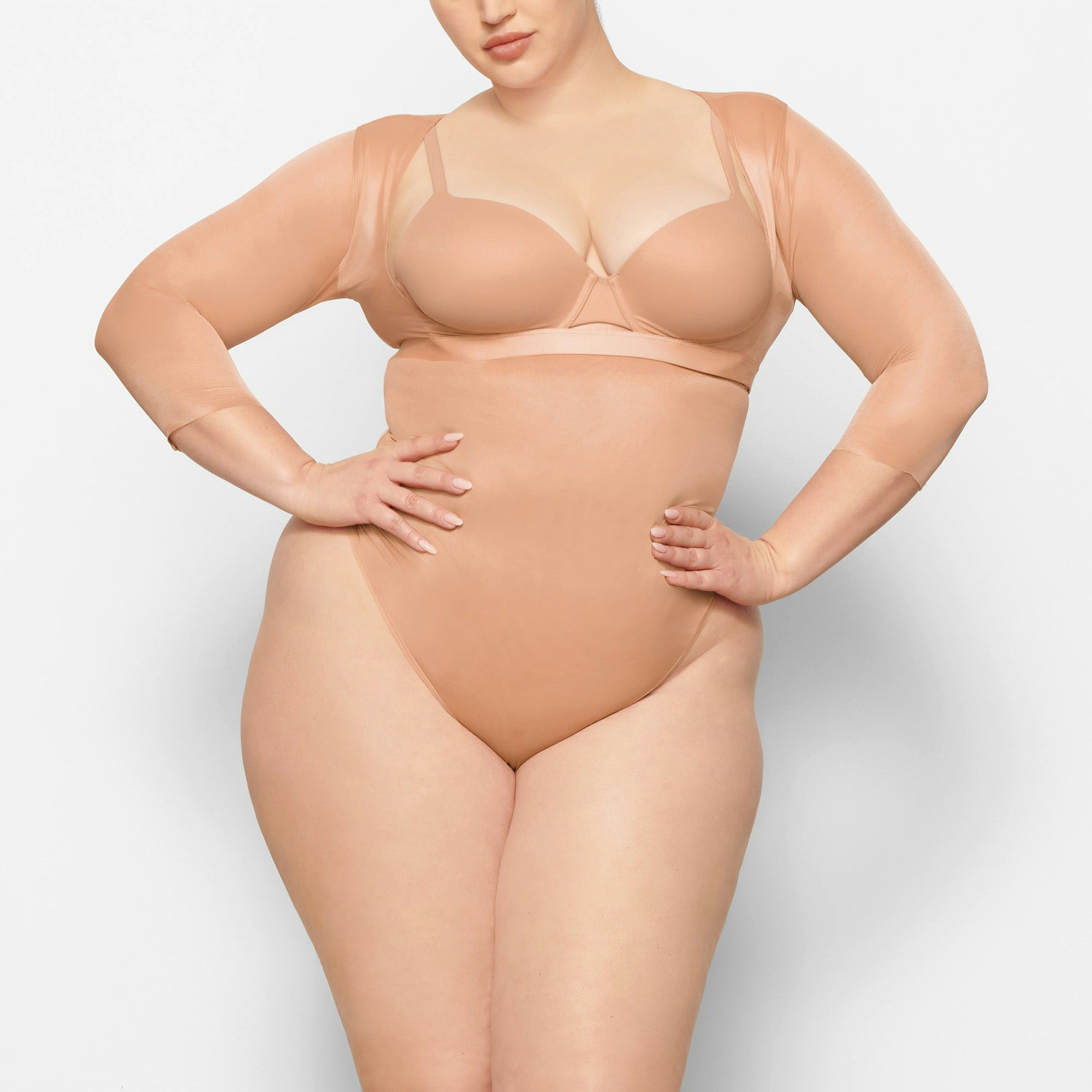 A MODEL STANDS FACING FORWARD WEARING THE SKIMS BARELY THERE OPEN BUST ARM SHAPER IN CLAY | SEE: 2X, 3X, 4X