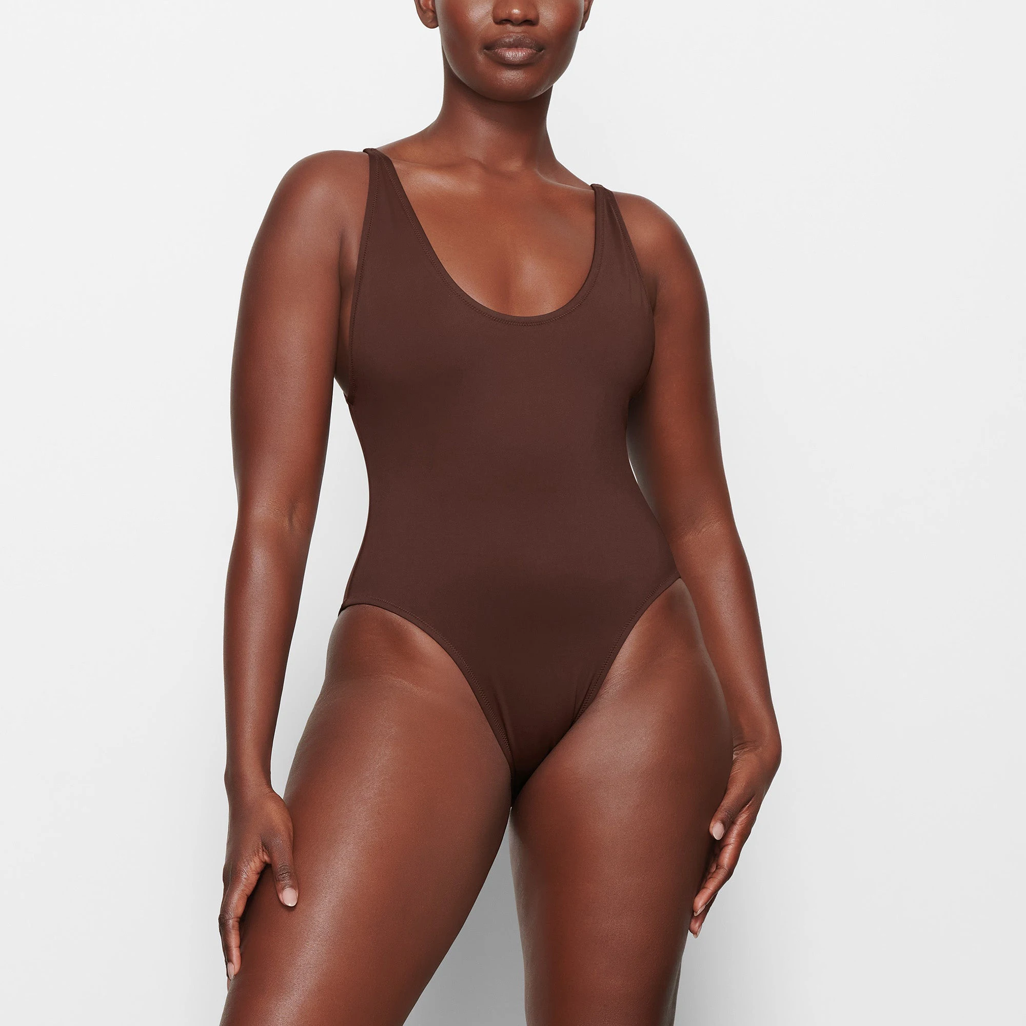 A MODEL STANDS FACING FORWARD WEARING THE SKIMS SWIM SCOOP NECK ONE PIECE IN COCOA | SEE: M, L, XL