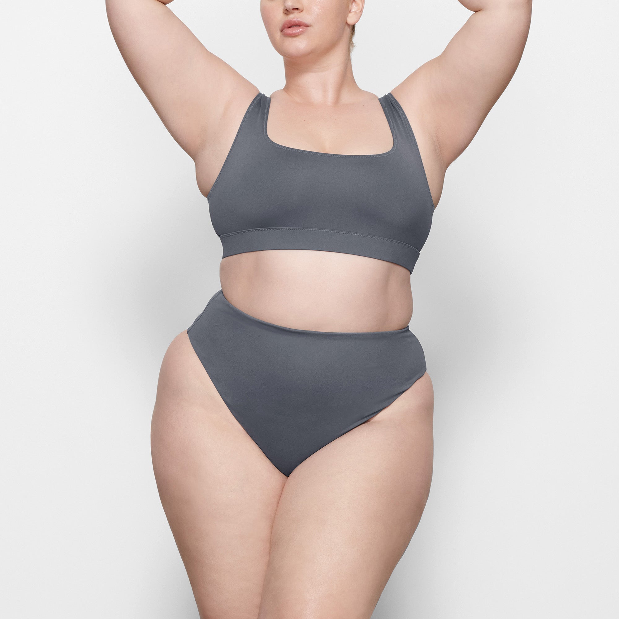 A MODEL STANDS FACING FORWARD WEARING THE SKIMS SWIM TANK BIKINI TOP IN GUNMETAL | SEE: 2X, 3X, 4X