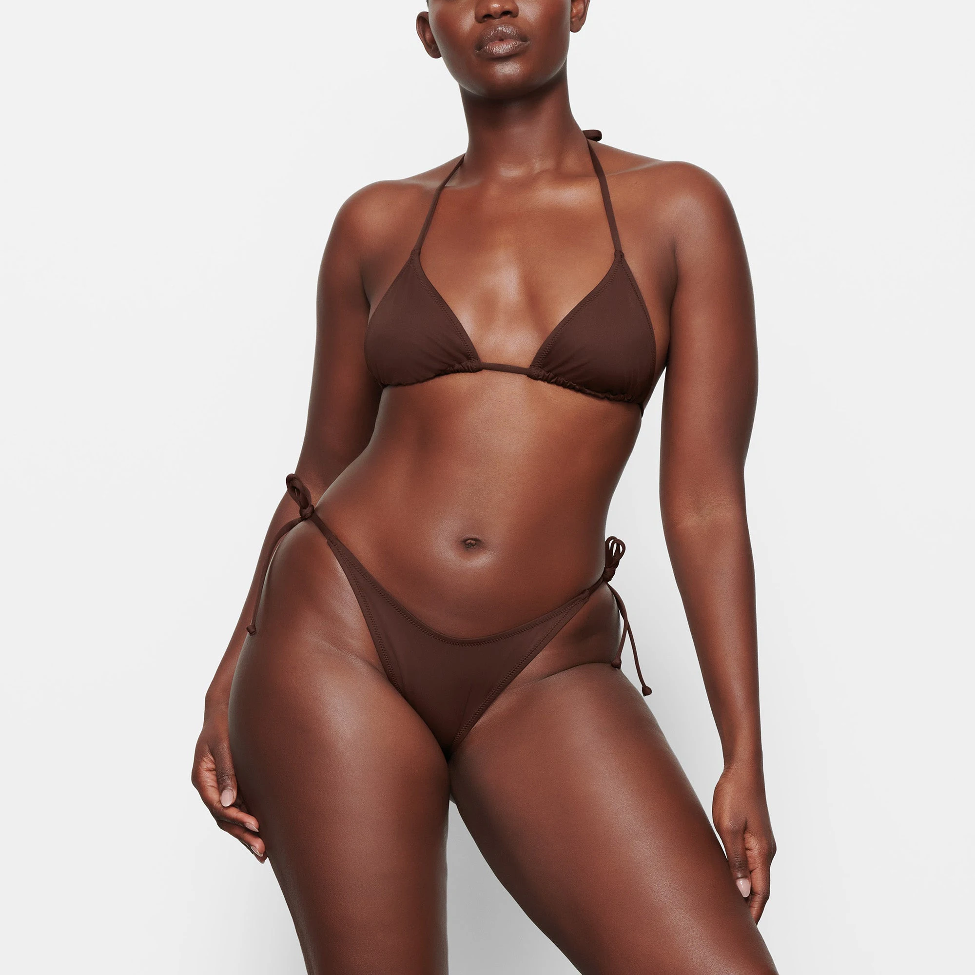 A MODEL STANDS FACING FORWARD WEARING THE SKIMS SWIM TRIANGLE TOP IN COCOA | SEE: M, L, XL